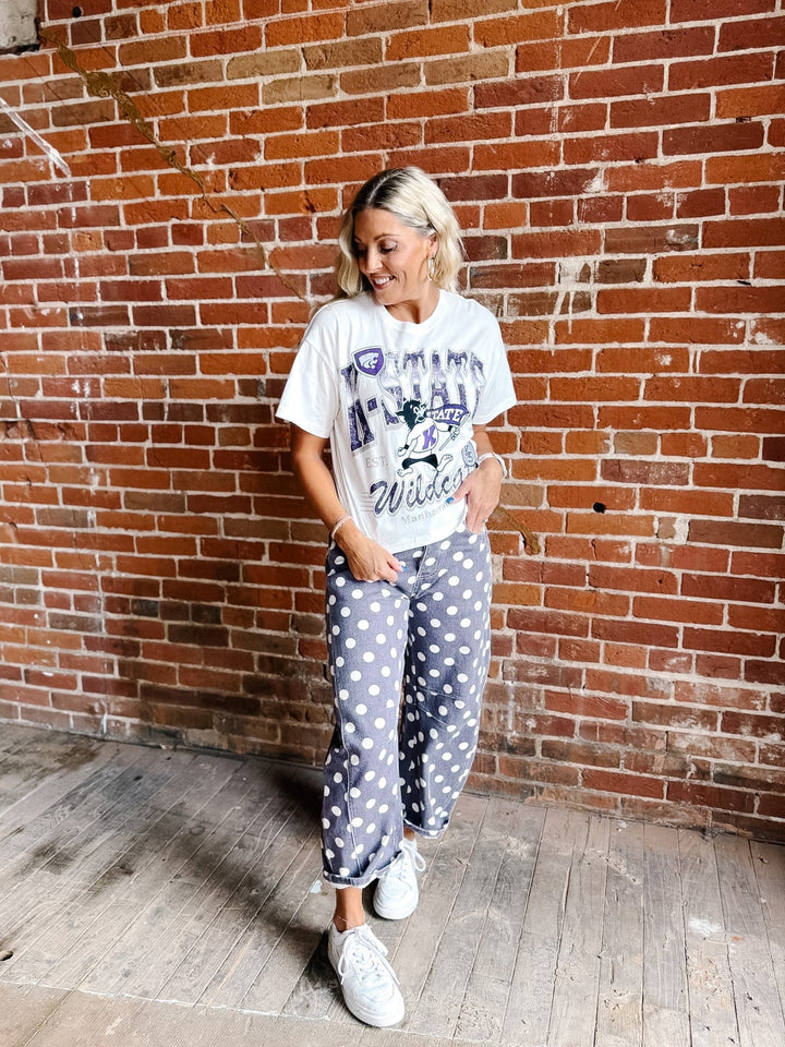 A woman stands in front of a red brick wall wearing a white K-State graphic t-shirt, gray pants with white polka dots, and white sneakers.