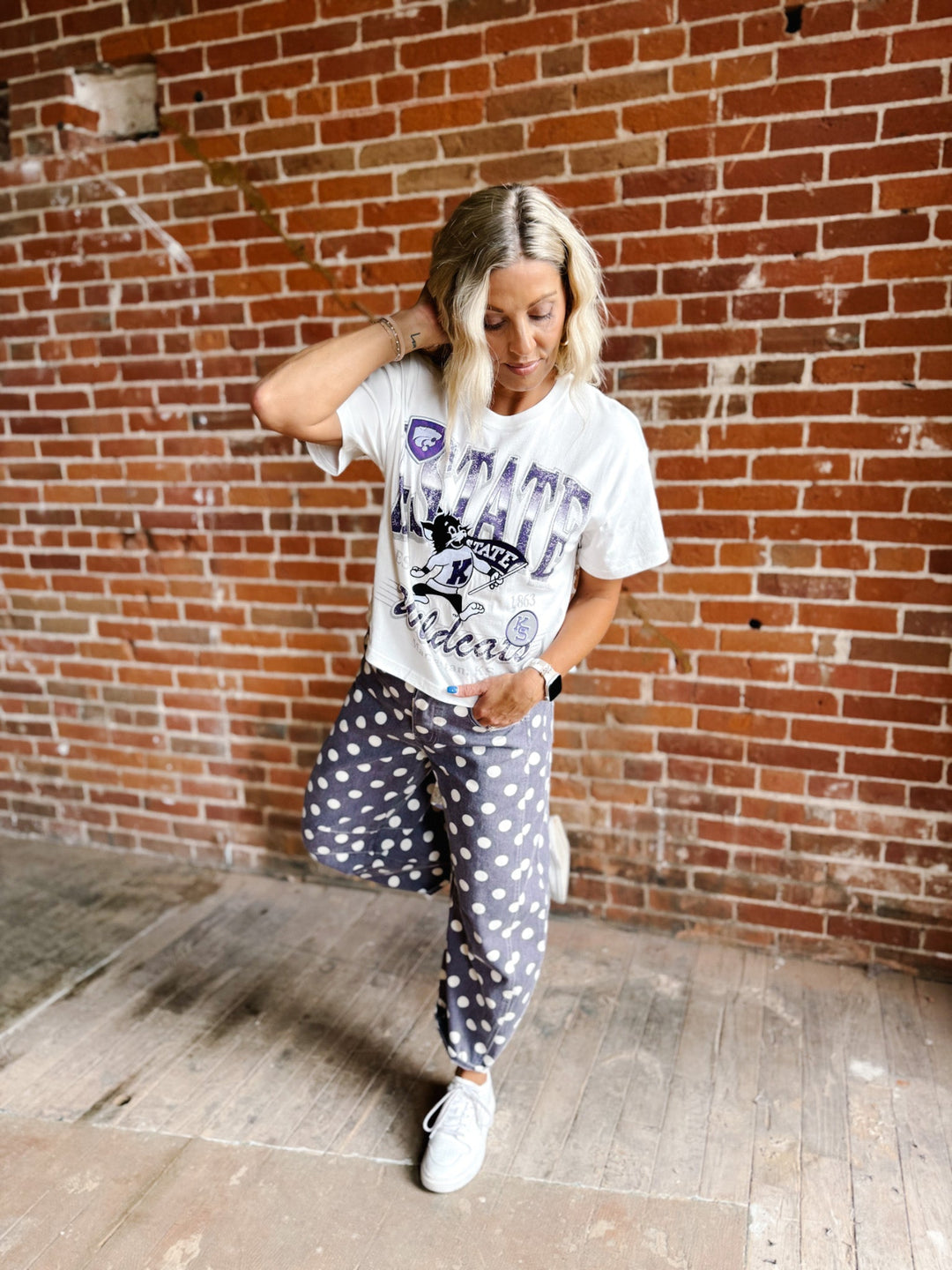 A woman posing in a white K-State graphic t-shirt and gray-and-white polka dot sweatpants against a red brick wall background.