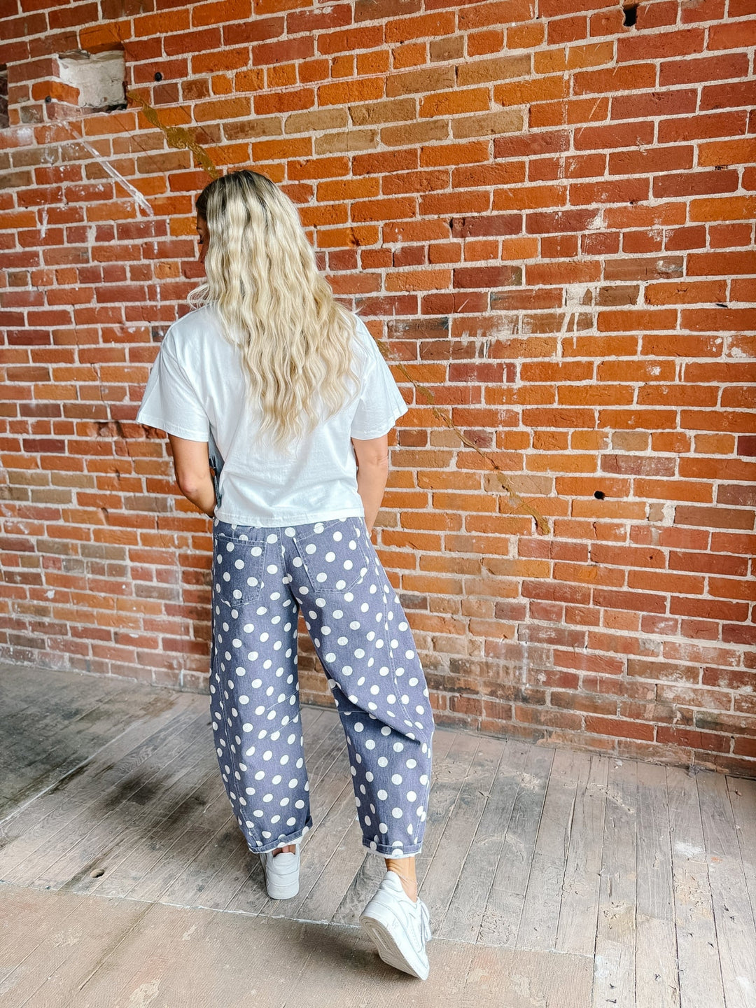 A woman with long blonde hair is seen from behind, wearing loose blue pants with white polka dots, a white t-shirt, and white sneakers, standing against a red brick wall.