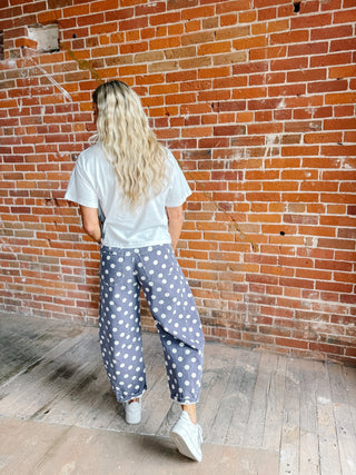 A woman with long blonde hair is seen from behind, wearing loose blue pants with white polka dots, a white t-shirt, and white sneakers, standing against a red brick wall.