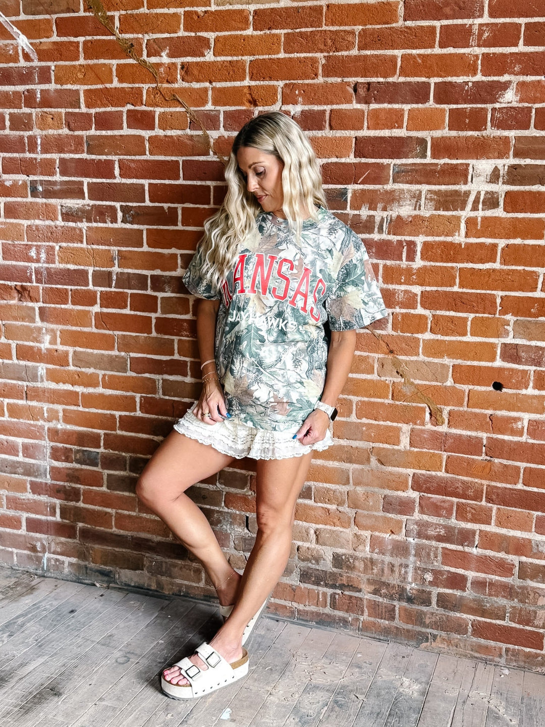 A woman posing against a red brick wall wearing a camouflage graphic t-shirt with &