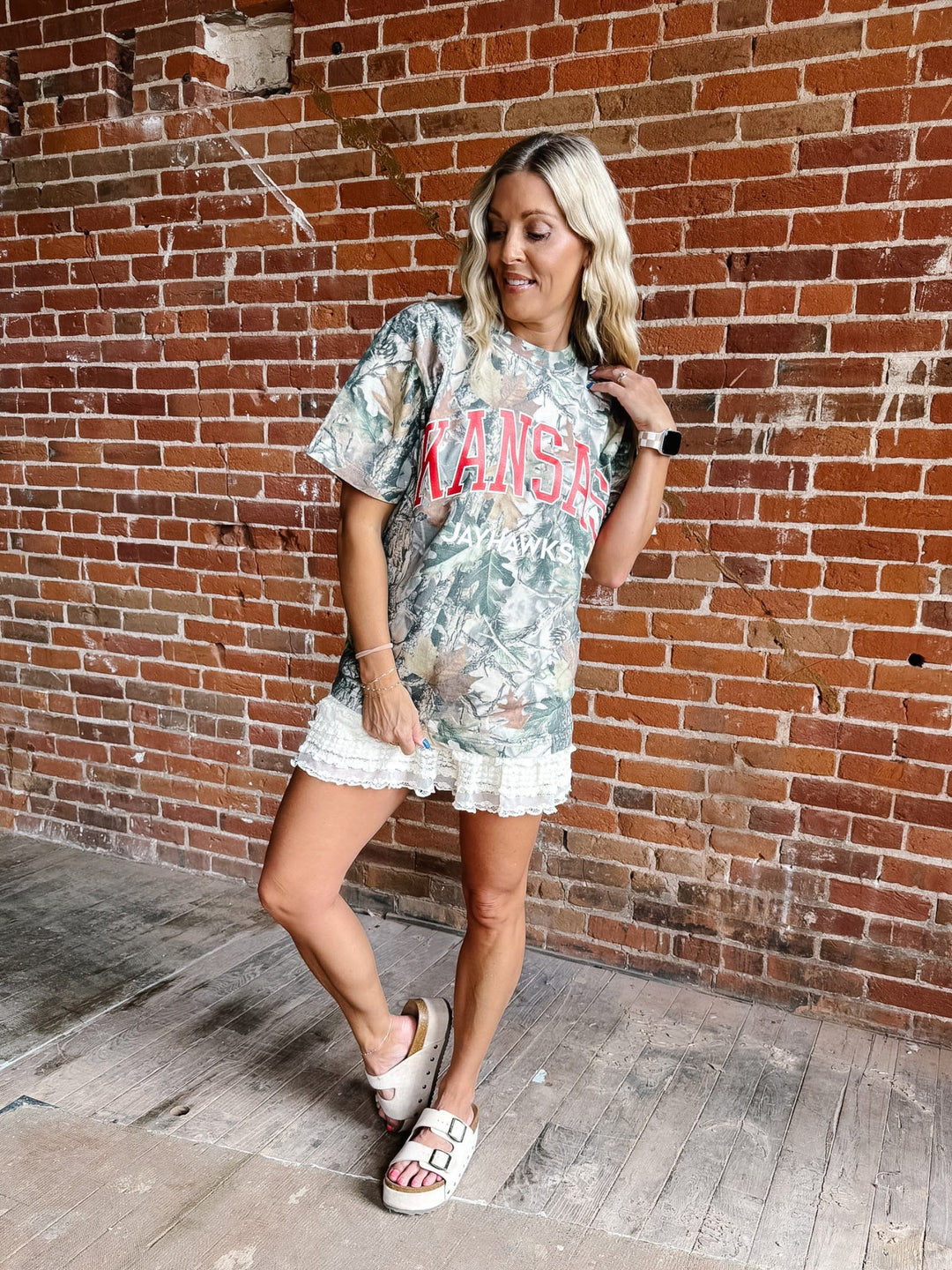 A woman posing in front of a brick wall wearing a Kansas Jayhawks graphic t-shirt, a white lace-trimmed mini skirt, and white slide sandals.