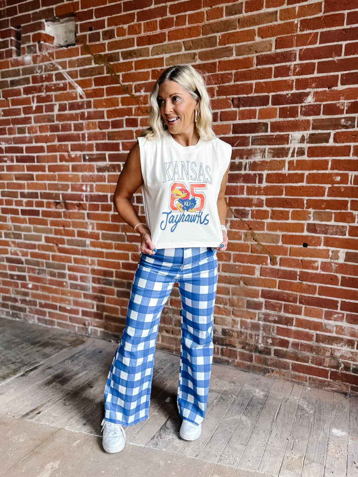 A woman stands in front of a red brick wall wearing a white sleeveless crop top with a blue and red &