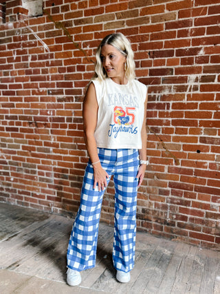 A woman stands in front of a red brick wall wearing a cream sleeveless crop top with a colorful 'Kansas Jayhawks 95' graphic, paired with blue and white gingham wide-leg pants and white sneakers.