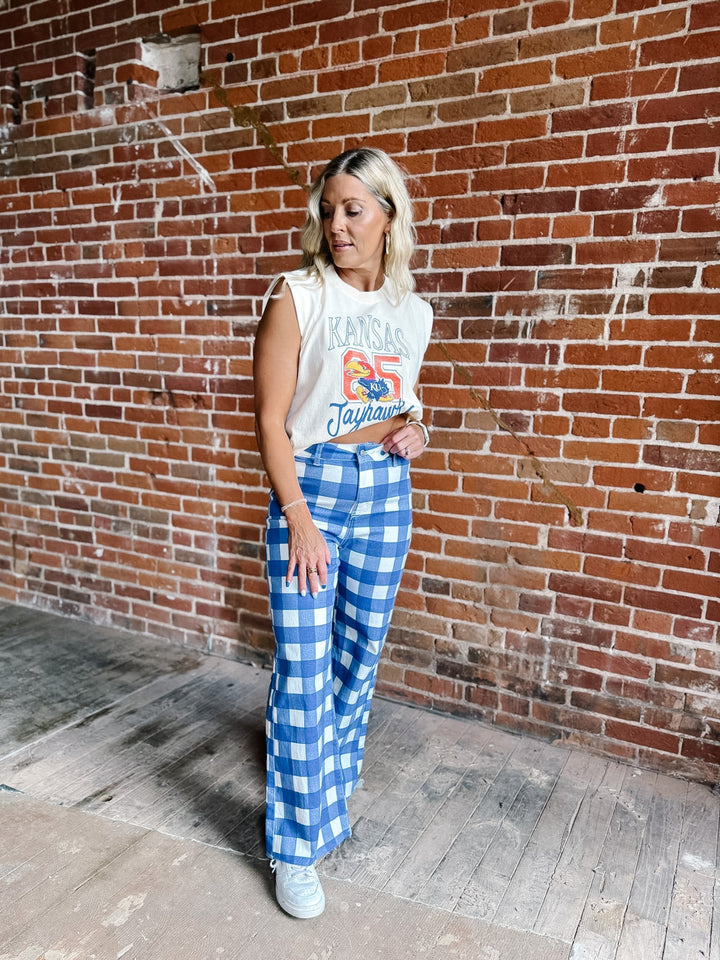 A woman stands against a red brick wall wearing a cream sleeveless graphic top and blue-and-white gingham wide-leg pants.