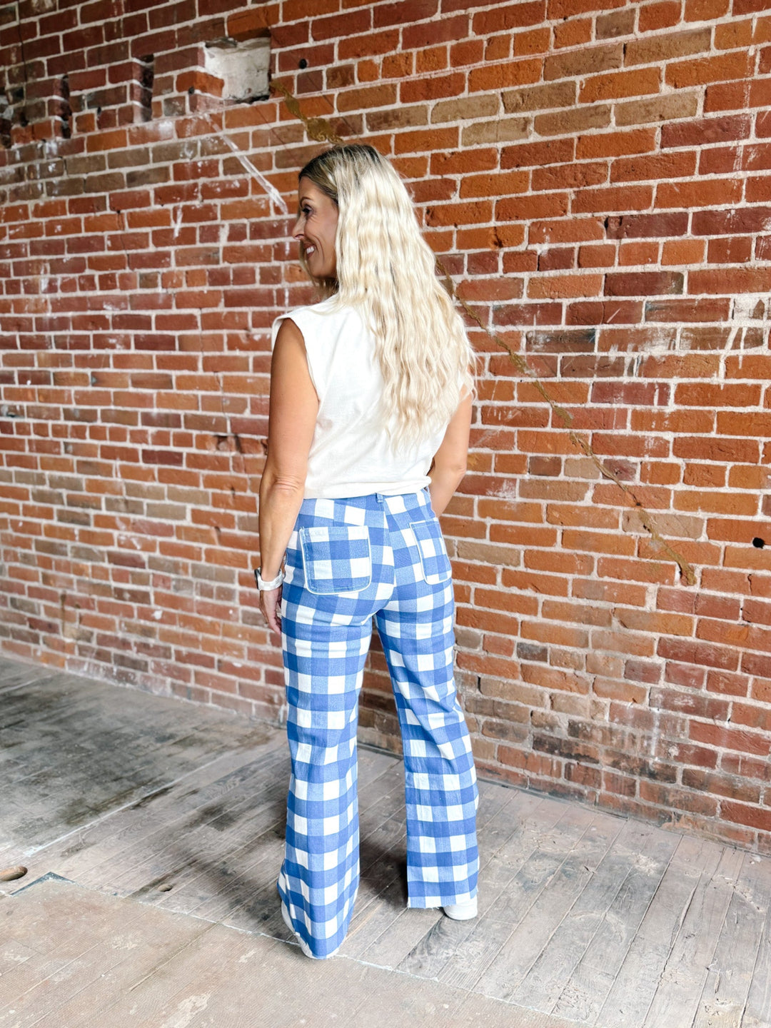 A woman with long blonde hair stands with her back to the camera, wearing blue and white gingham wide-leg pants and a white sleeveless top, posing in front of a red brick wall.