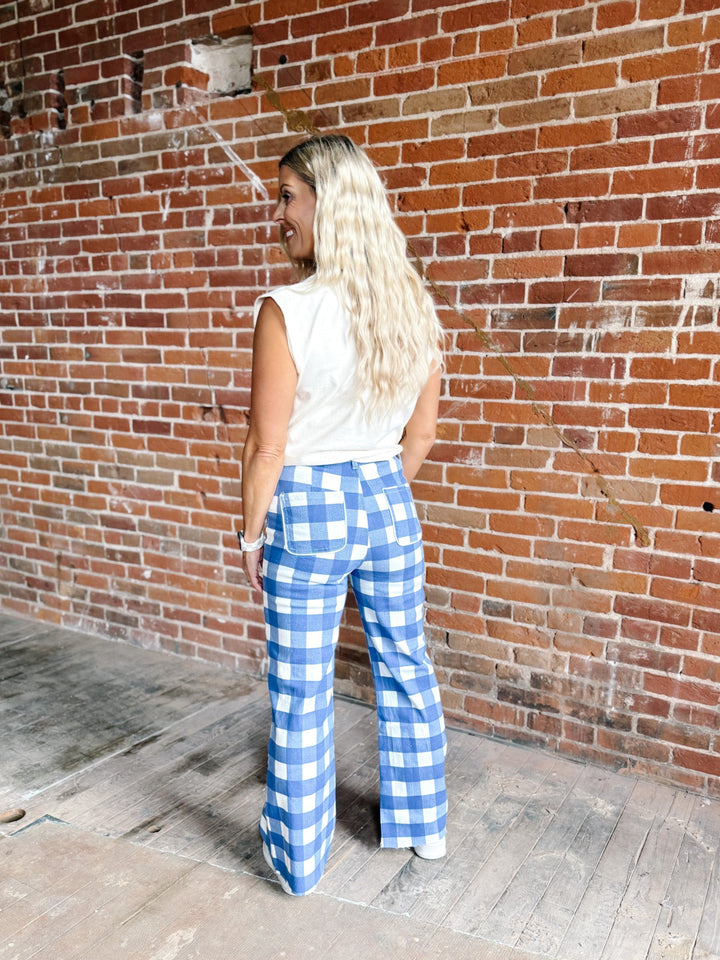 A woman with long blonde hair stands with her back to the camera, wearing blue and white gingham wide-leg pants and a white sleeveless top, posing in front of a red brick wall.