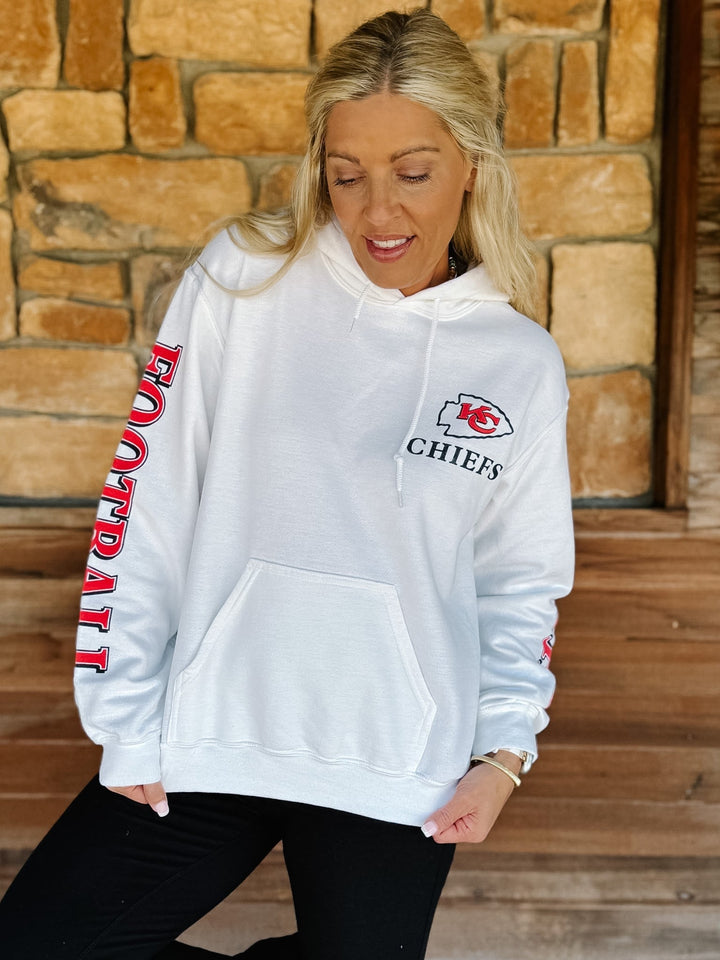 A woman wearing a white Kansas City Chiefs football hoodie with red and black graphics, posing in front of a rustic stone wall.