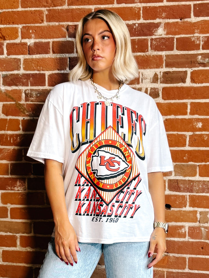 A woman with blonde hair stands against a red brick wall wearing a white Kansas City Chiefs graphic t-shirt and light blue jeans.