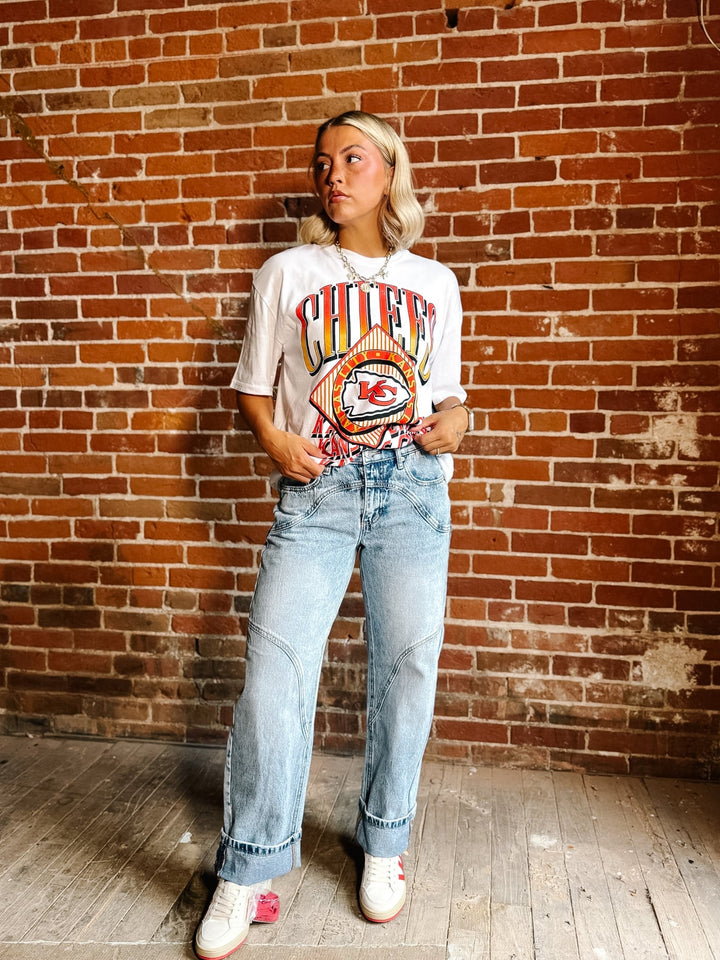 A woman stands against a red brick wall wearing a white graphic t-shirt with a colorful &