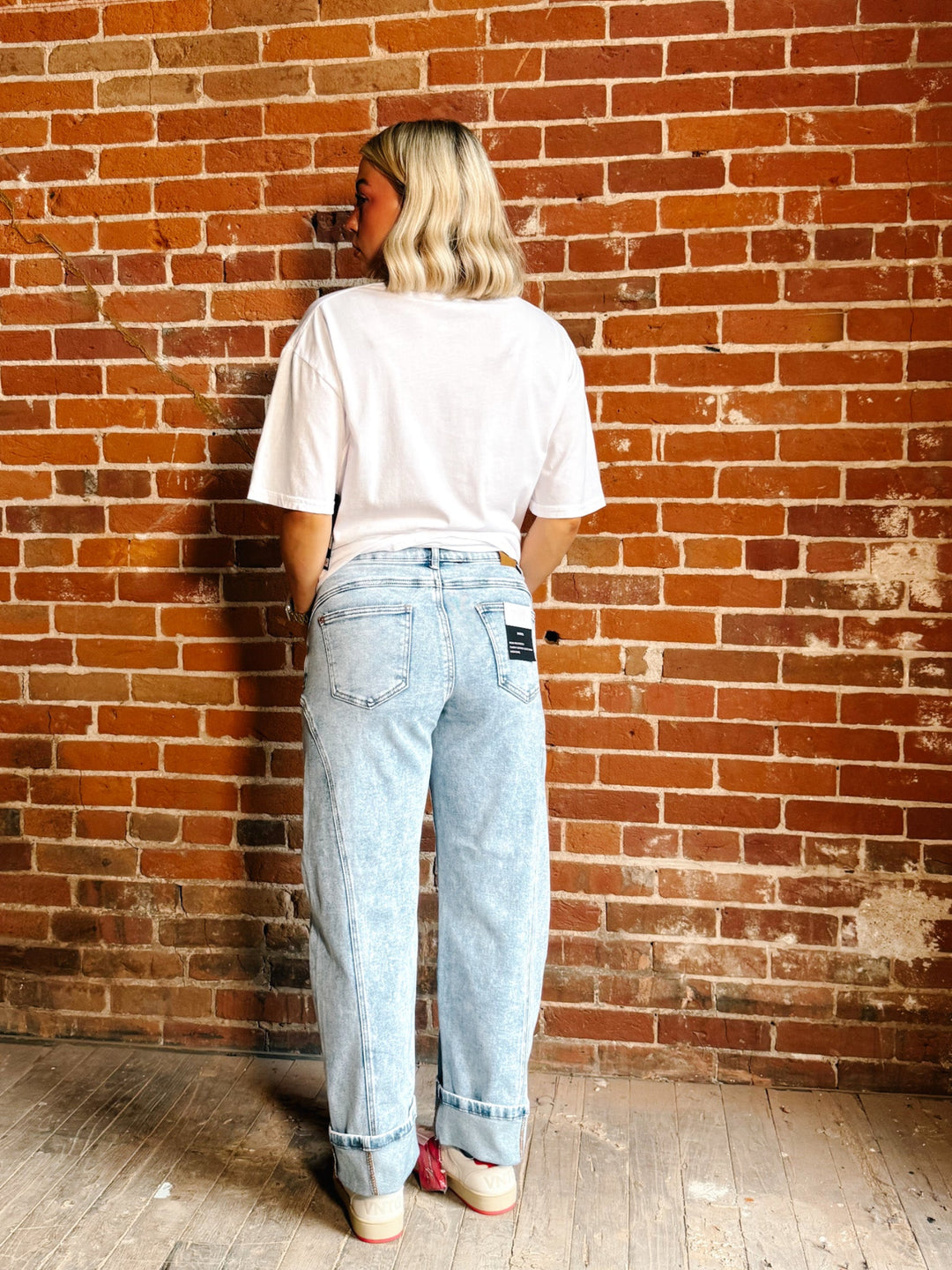 A person standing with their back to the camera, wearing light-wash blue jeans, a white t-shirt, and pink sneakers, set against a red-brown brick wall background.