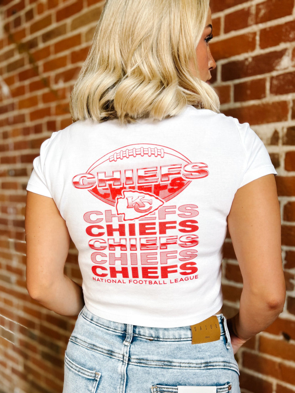 A woman viewed from behind wearing a white t-shirt with a large red Kansas City Chiefs graphic on the back, paired with light wash blue jeans, standing against a red brick wall.