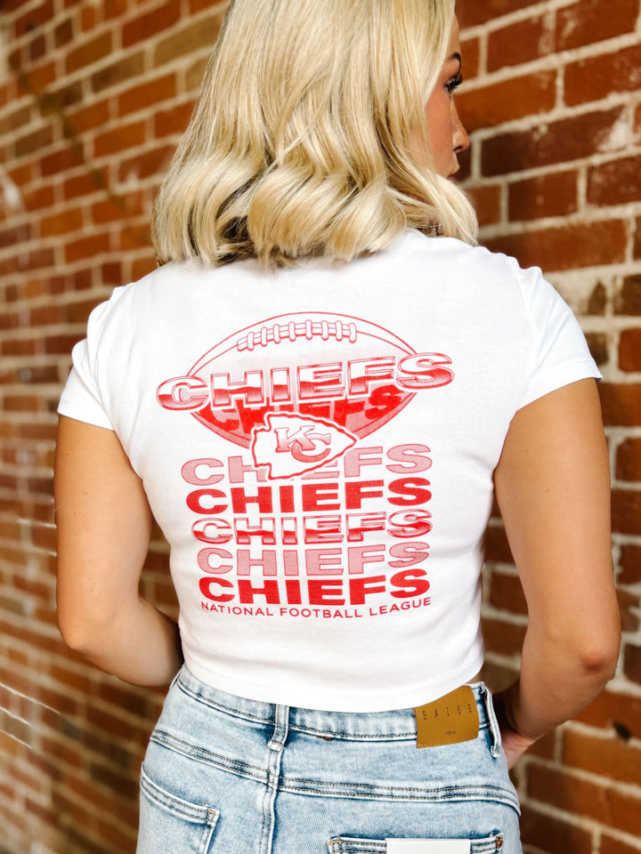 A woman viewed from behind wearing a white t-shirt with a large red Kansas City Chiefs graphic on the back, paired with light wash blue jeans, standing against a red brick wall.