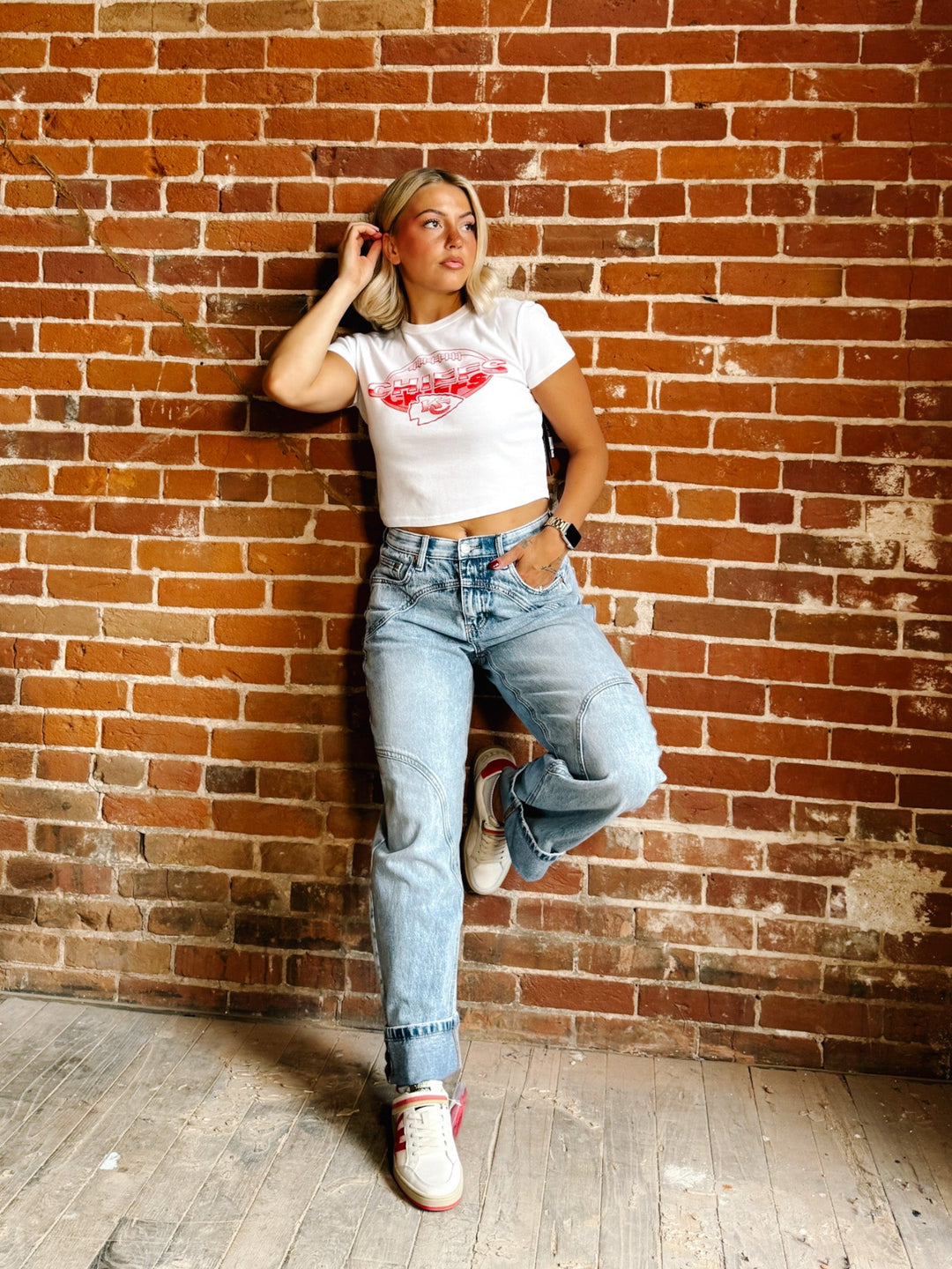 A woman posing in a white graphic crop top, light-wash blue jeans, and white sneakers against a red-brown brick wall.