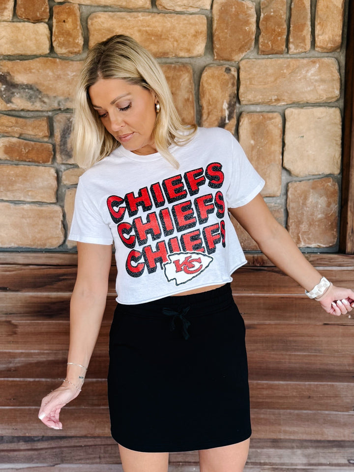 A woman wearing a white Kansas City Chiefs cropped graphic t-shirt with red and black lettering, paired with a black mini skirt, standing in front of a rustic stone wall.