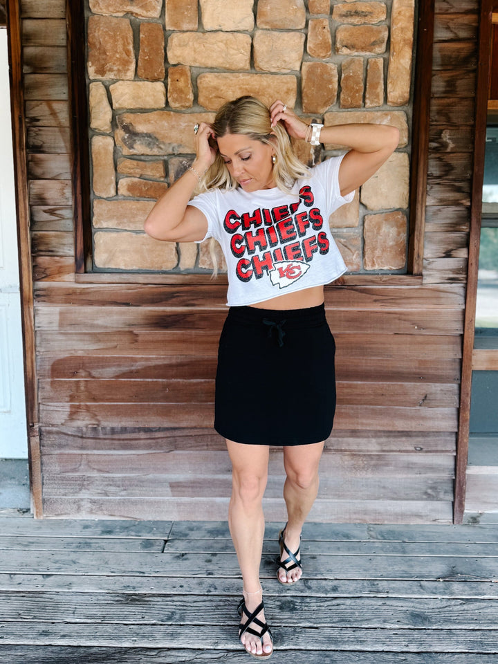 A woman stands on a rustic wooden porch wearing a white cropped graphic T-shirt with &