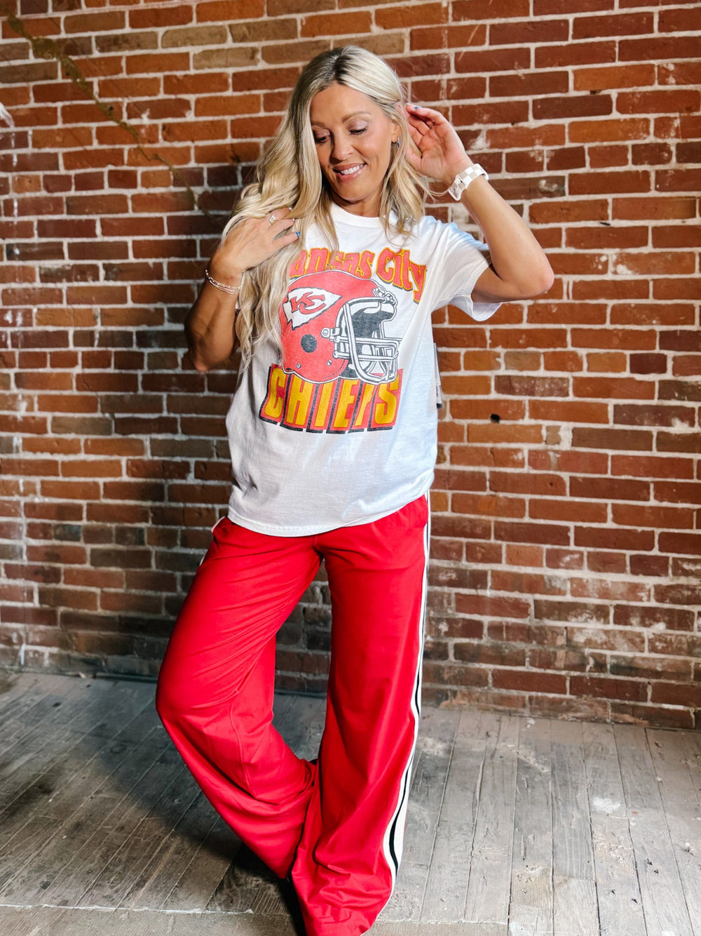 A woman posing in front of a brick wall wearing a white graphic t-shirt with a red and orange football helmet design and the words 'CHICAGO CHILIES', paired with bright red track pants featuring a white and black side stripe.
