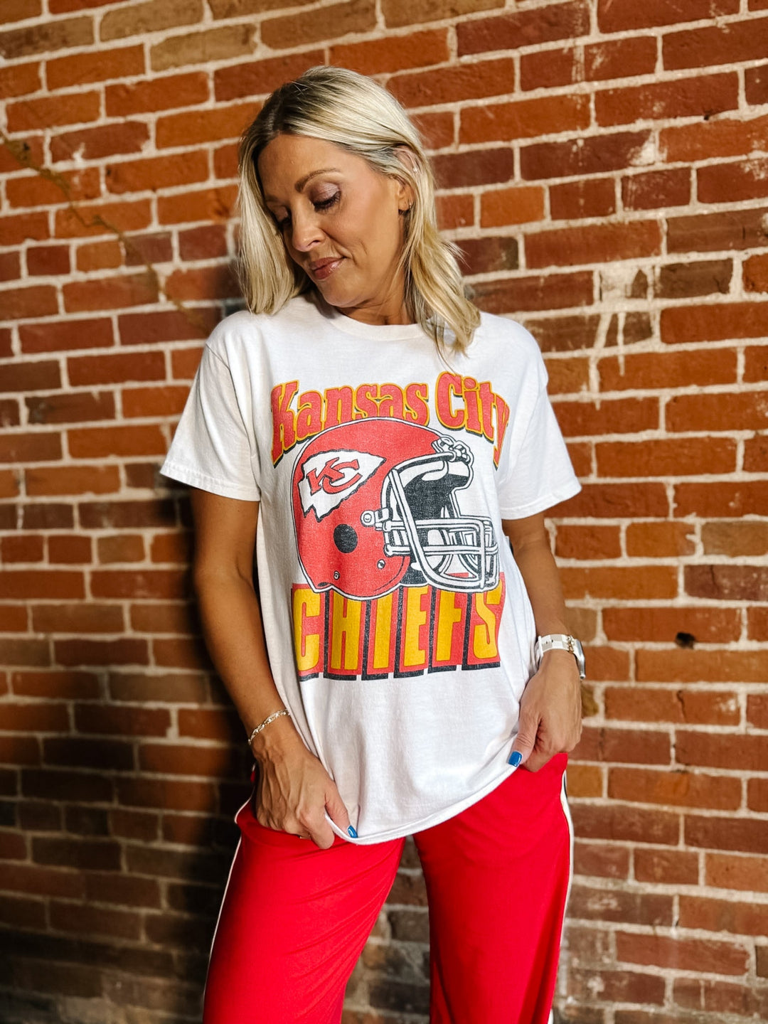 A woman wearing a white Kansas City Chiefs graphic t-shirt and red sweatpants, standing against a red-brown brick wall.
