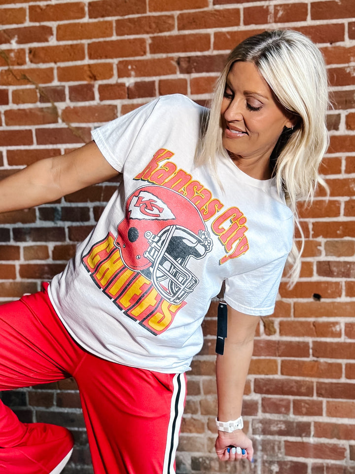 A woman posing in front of a brick wall wearing a white Kansas City Chiefs graphic t-shirt and red track pants.