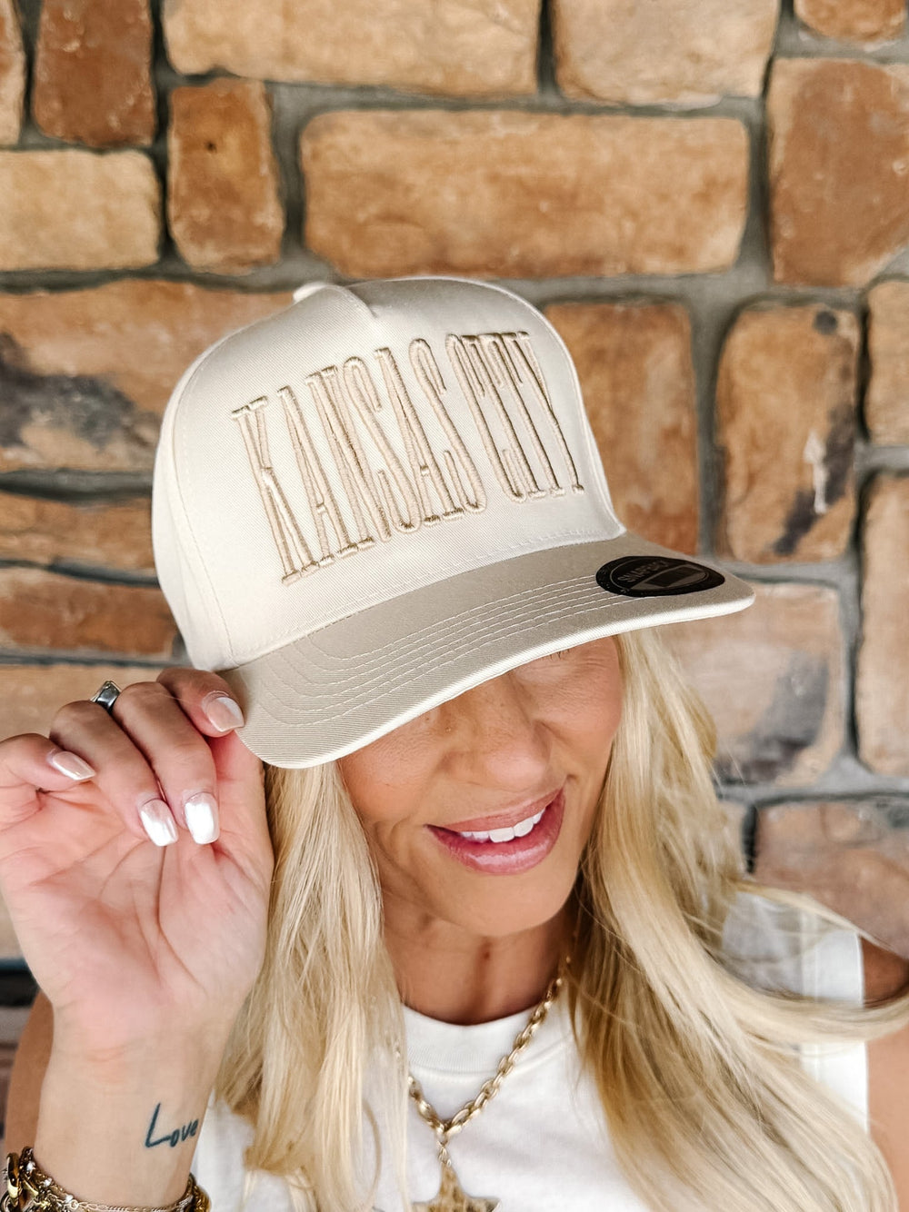 A woman wearing a cream-colored snapback cap with 'Kansas City' embroidered in gold across the front, posing in front of a rustic stone wall.