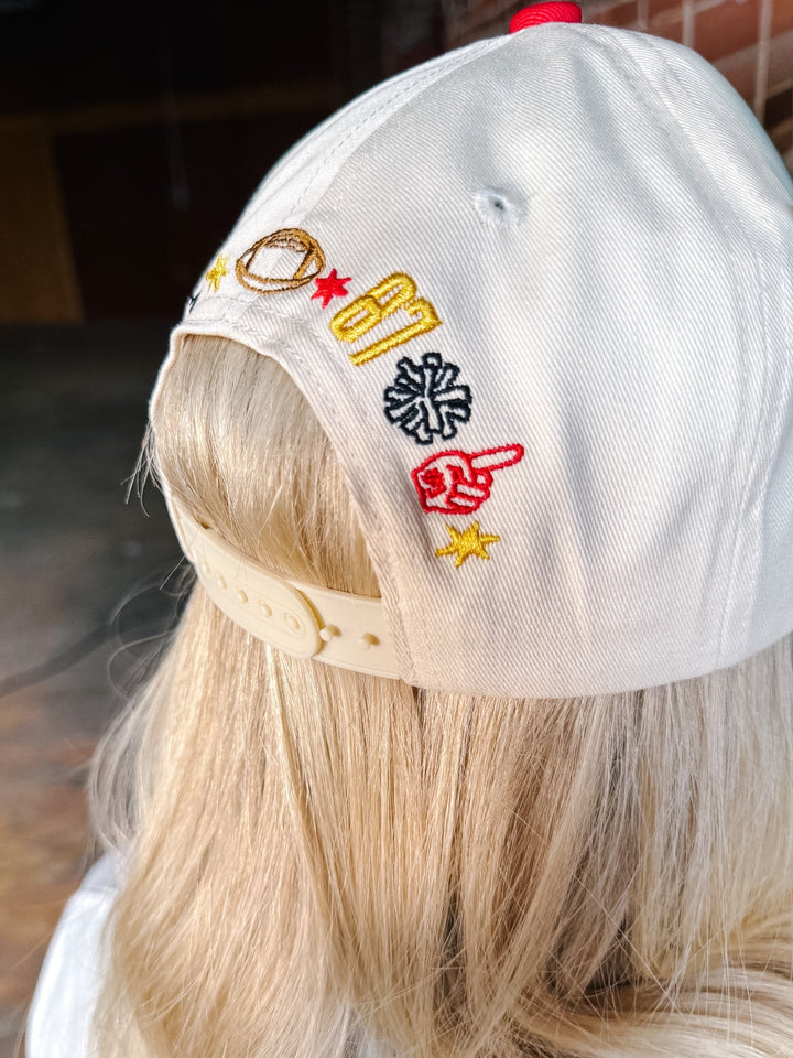 A close-up back view of a person wearing a white baseball cap with colorful embroidered lettering and symbols on the side, including yellow and red stars and a red pointing hand.