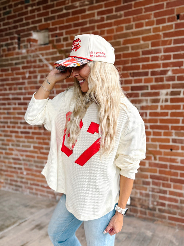 A woman wearing a cream waffle-knit sweatshirt with a large red graphic, light blue jeans, and a cream baseball cap with red text, standing in front of a red brick wall.