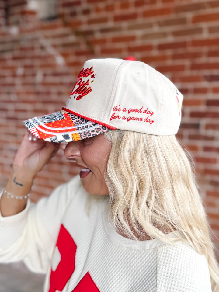 A woman wearing a white baseball cap with red embroidered text and a multicolor patterned brim, standing in front of a brick wall.