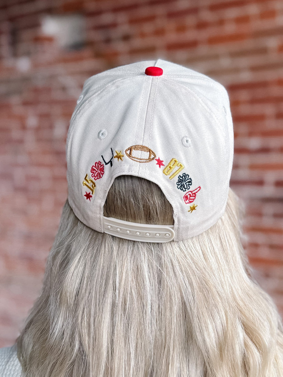 A white baseball cap viewed from the back, worn by a person with long blonde hair. The cap features colorful embroidered sports-themed motifs including a football, basketball, baseball, and stars, with a red button on top and an adjustable strap closure.