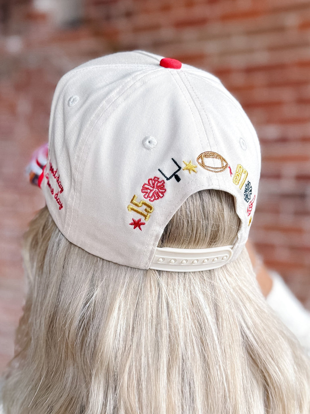 A person viewed from behind wearing a white baseball cap with colorful embroidered letters and symbols on the back, set against a blurred brick wall background.