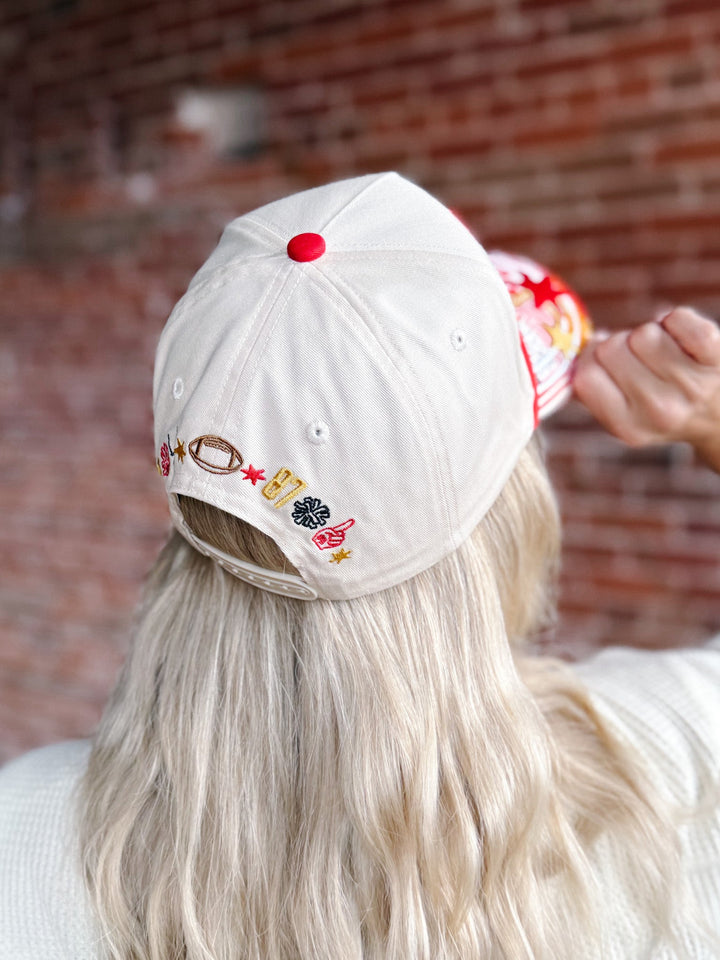 A person viewed from behind wearing a cream-colored baseball cap with embroidered details and a red button on the crown, set against a blurred brick wall background.