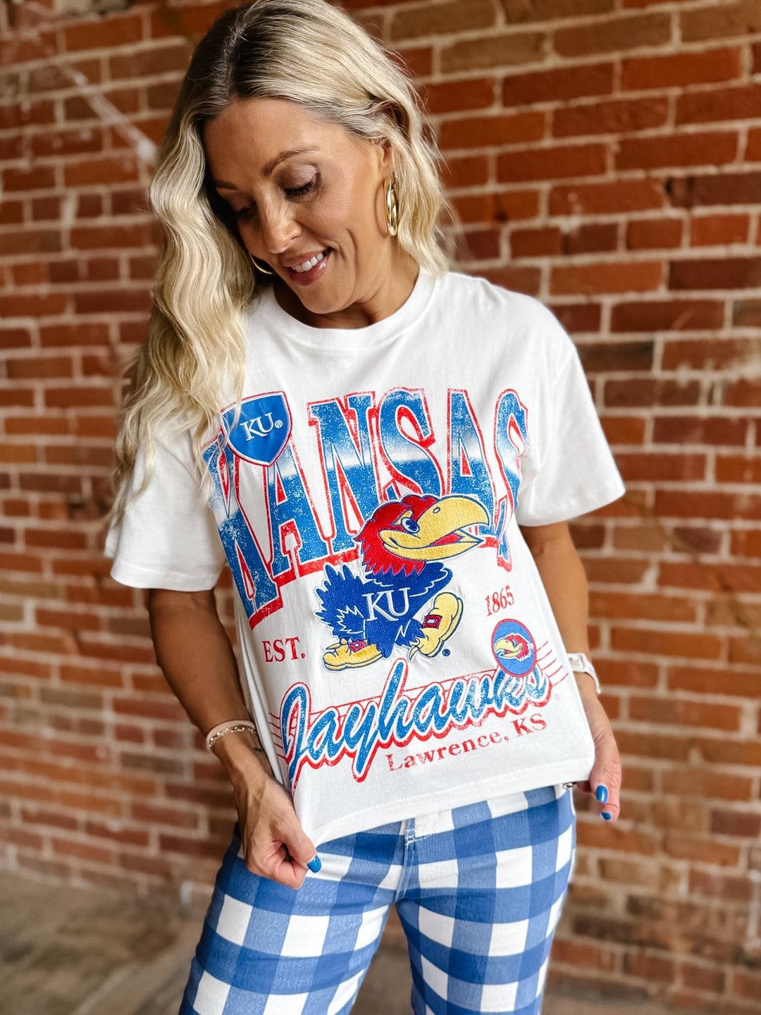 A woman wearing a white Kansas Jayhawks graphic t-shirt and blue and white gingham pants, standing in front of a red brick wall.