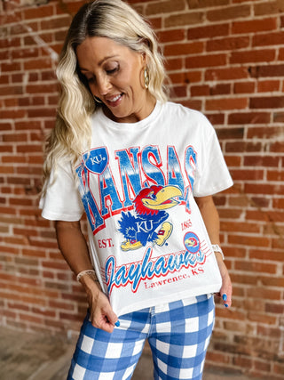A woman wearing a white Kansas Jayhawks graphic t-shirt and blue and white gingham pants, standing in front of a red brick wall.