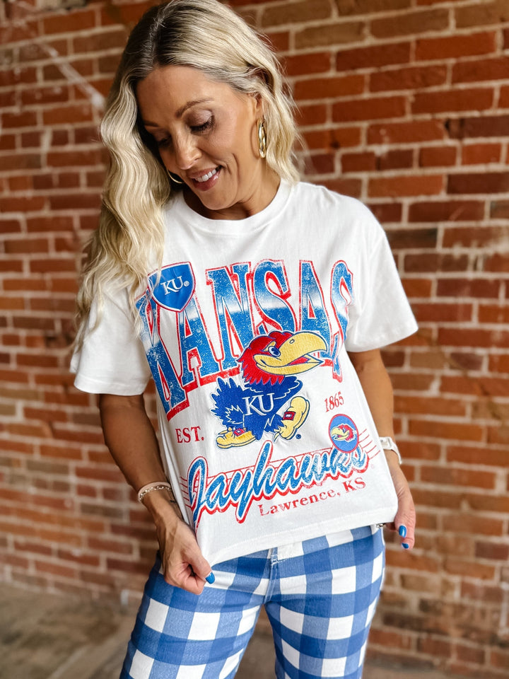 A woman wearing a white Kansas Jayhawks graphic t-shirt and blue and white gingham pants, standing in front of a red brick wall.