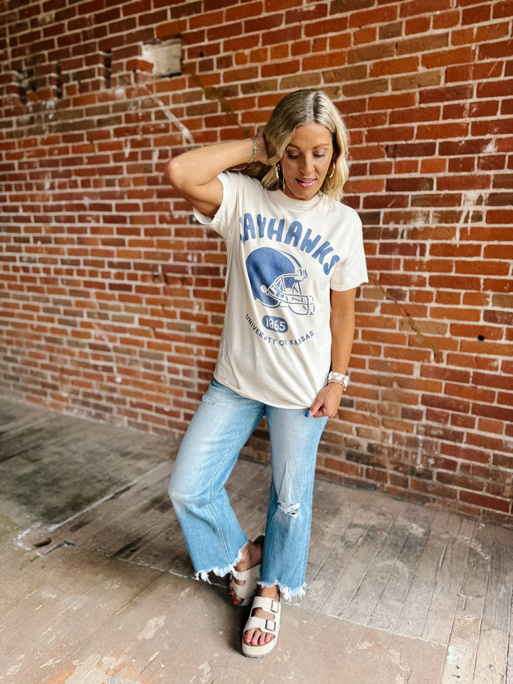 A woman posing in front of a brick wall wearing a cream-colored graphic t-shirt with a blue &