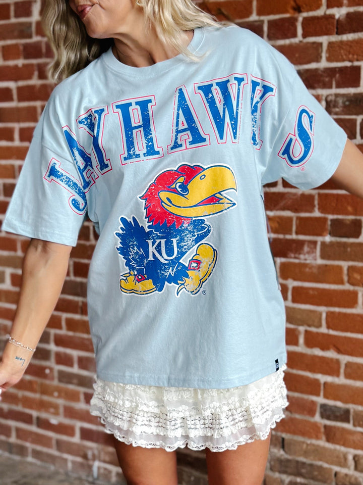 A woman wearing a light blue Kansas Jayhawks graphic t-shirt with a large distressed blue, red, and yellow jayhawk logo on the front, paired with a white lace hem skirt, standing against a red brick wall.