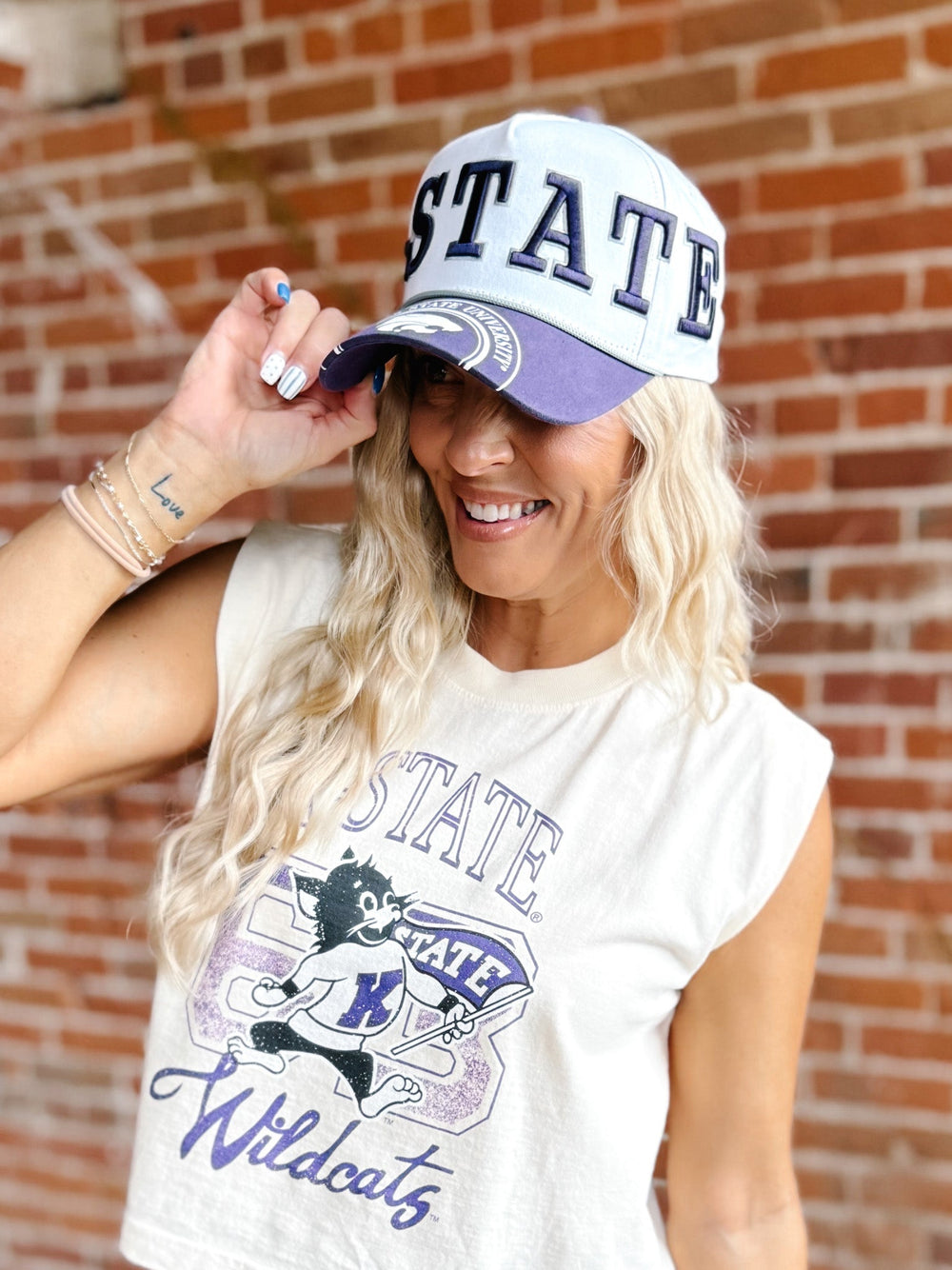 A woman wearing a white and purple baseball cap with 'STATE' and a wildcat graphic, paired with a matching white sleeveless graphic t-shirt, posing in front of a brick wall.