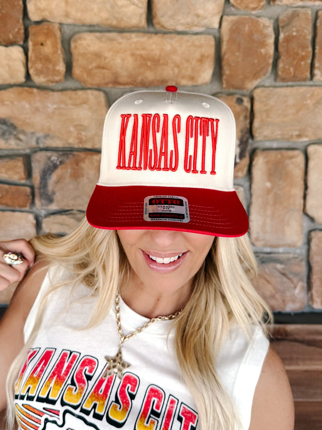 A woman wearing a red and white snapback cap with 'KANSAS CITY' in red letters on the front, posed in front of a rustic stone wall.
