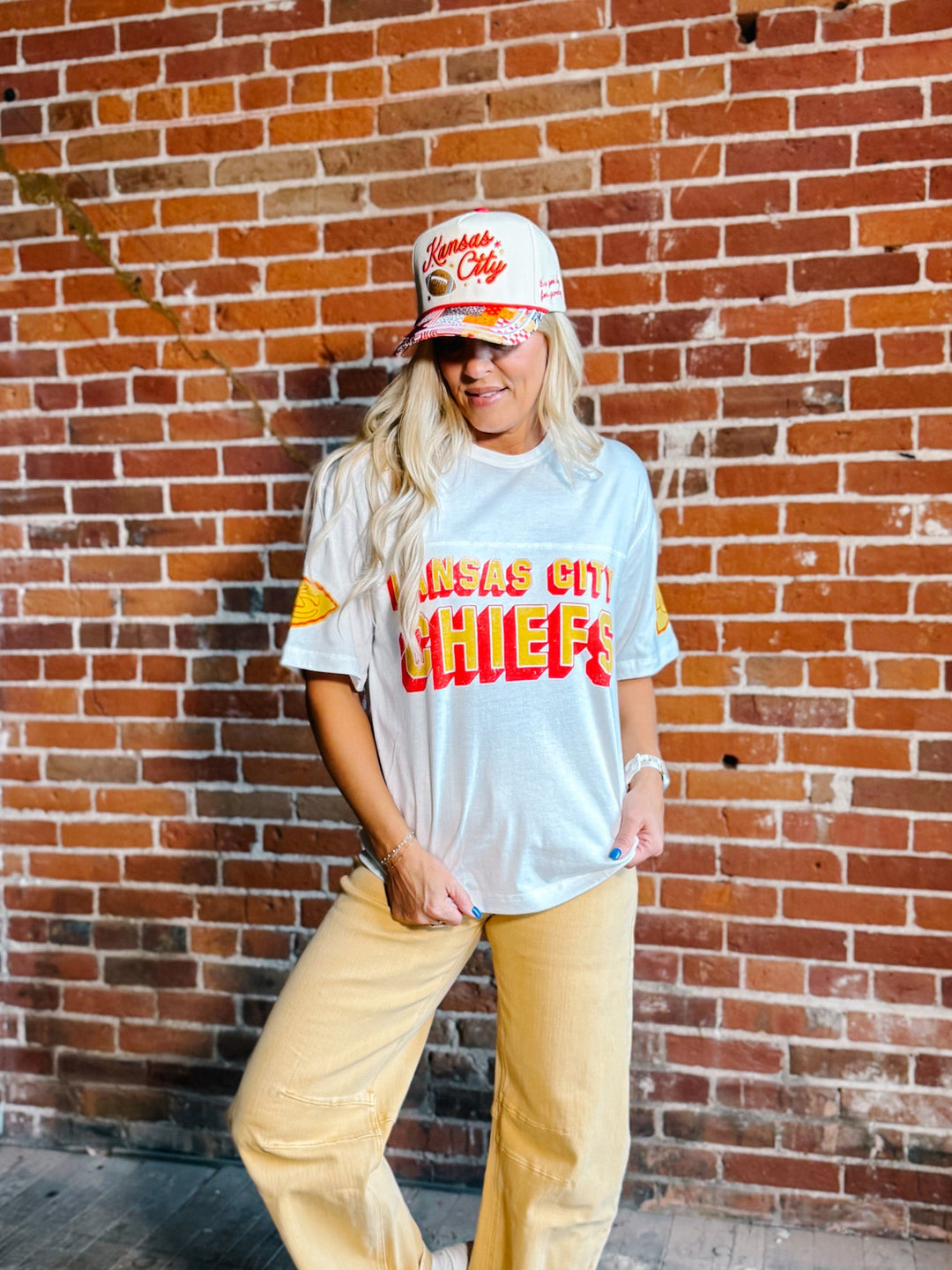 A woman wearing a light blue Kansas City Chiefs graphic t-shirt, matching yellow pants, and a white Chiefs cap stands against a red brick wall.