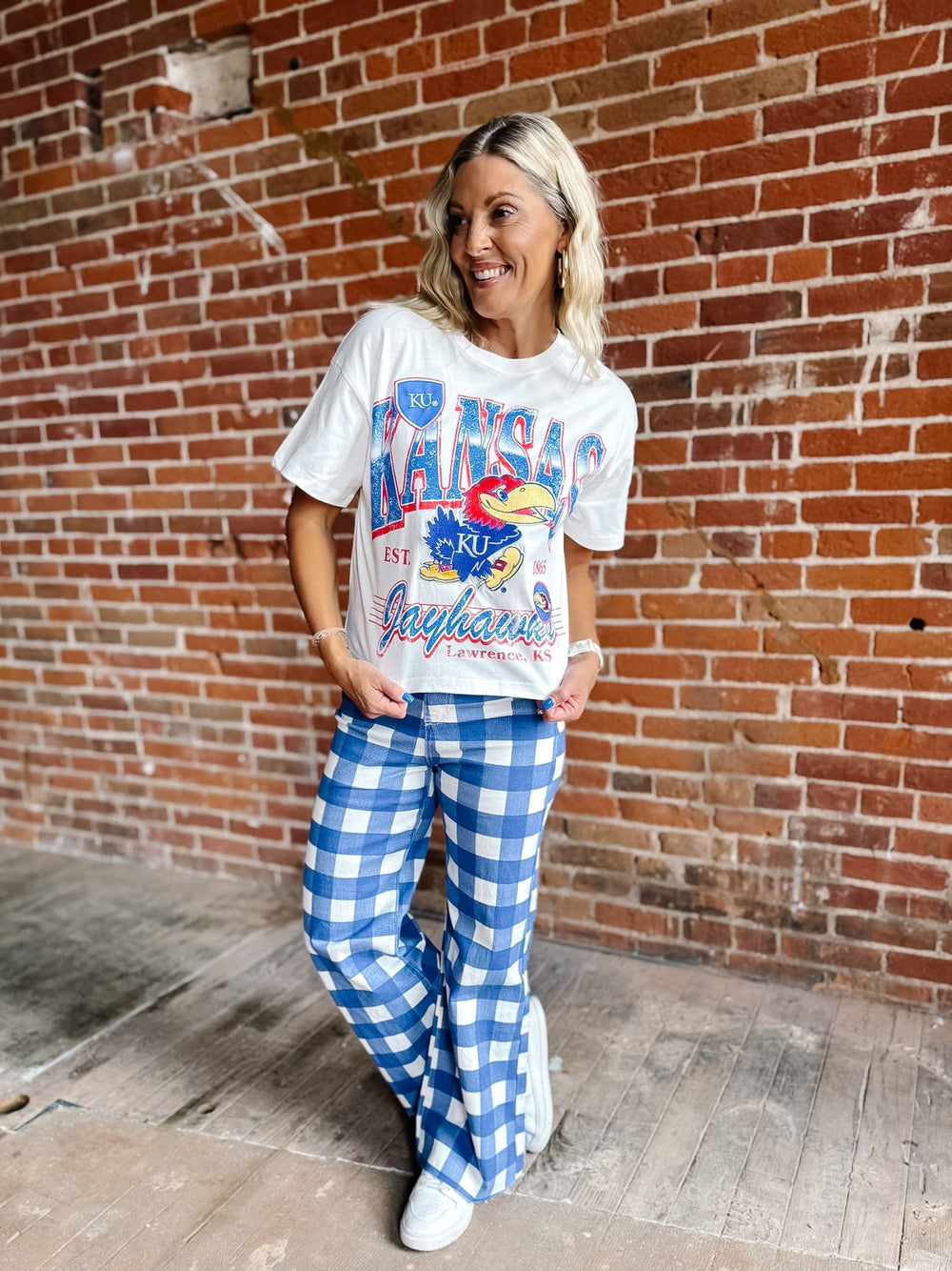 A woman posing in front of a red brick wall, wearing a white graphic t-shirt with blue and red 'Kansas Jayhawks' lettering and blue-and-white gingham checkered pants.