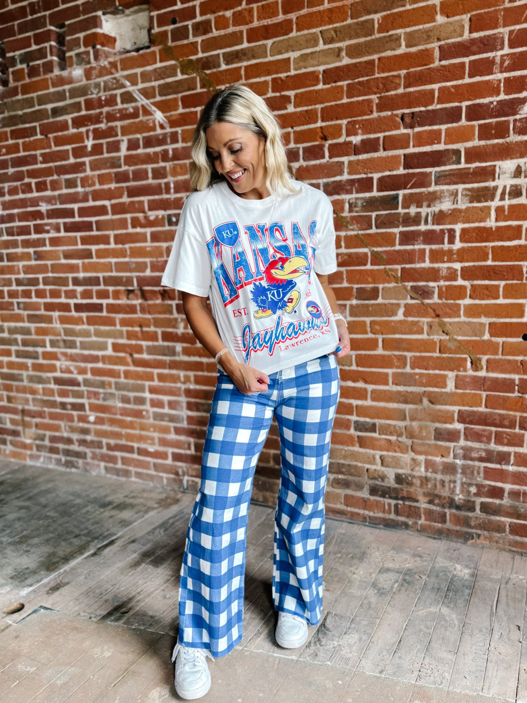 A woman stands in front of a red brick wall wearing a white graphic t-shirt with a blue and red Jayhawks print, paired with blue and white gingham wide-leg pants and white sneakers.