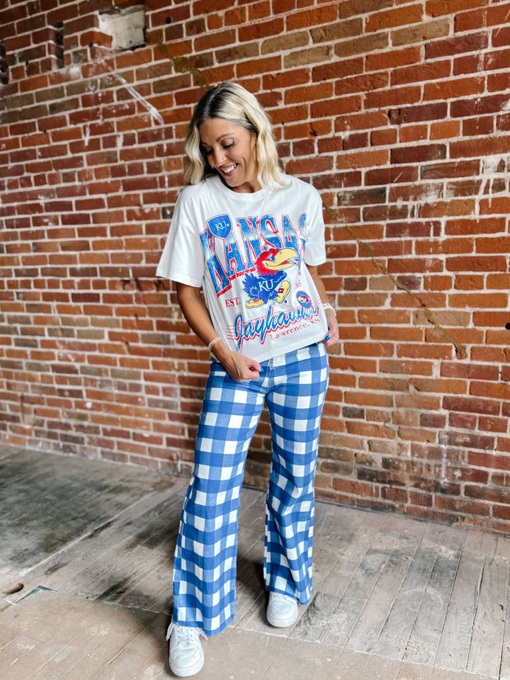 A woman stands in front of a red brick wall wearing a white graphic t-shirt with a blue and red Jayhawks print, paired with blue and white gingham wide-leg pants and white sneakers.
