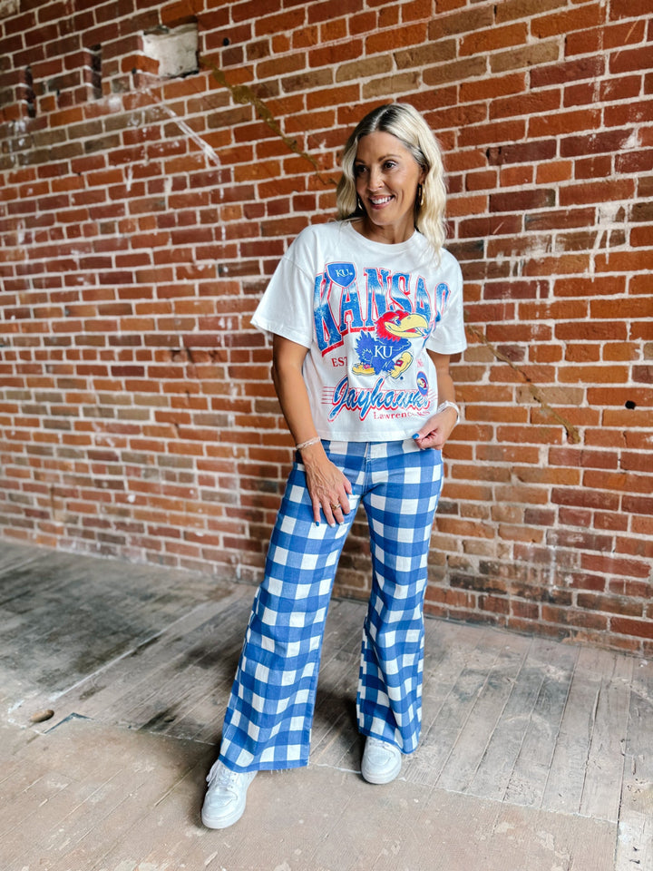 A woman stands in front of a red brick wall wearing a white graphic t-shirt and blue-and-white gingham wide-leg pants, paired with white sneakers.