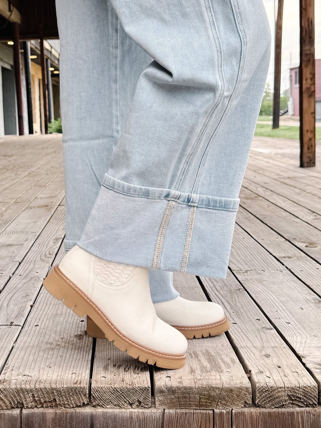 A person wearing light blue jeans and off-white ankle boots with tan soles, standing on a weathered wooden deck.