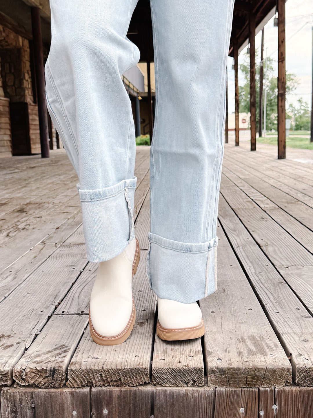 A person wearing light-wash blue jeans and off-white ankle boots with tan soles, standing on a weathered wooden boardwalk.
