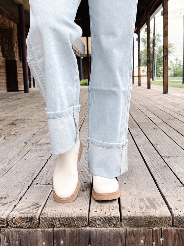 A person wearing light-wash blue jeans and off-white ankle boots with tan soles, standing on a weathered wooden boardwalk.