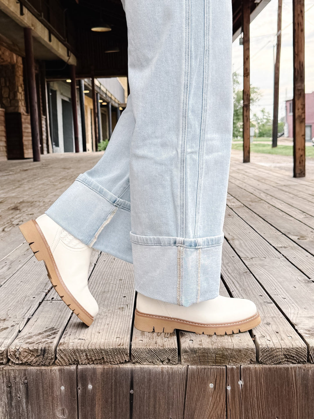 A person wearing light blue cuffed jeans and off-white ankle boots with tan soles, standing on a rustic wooden deck beside a porch.