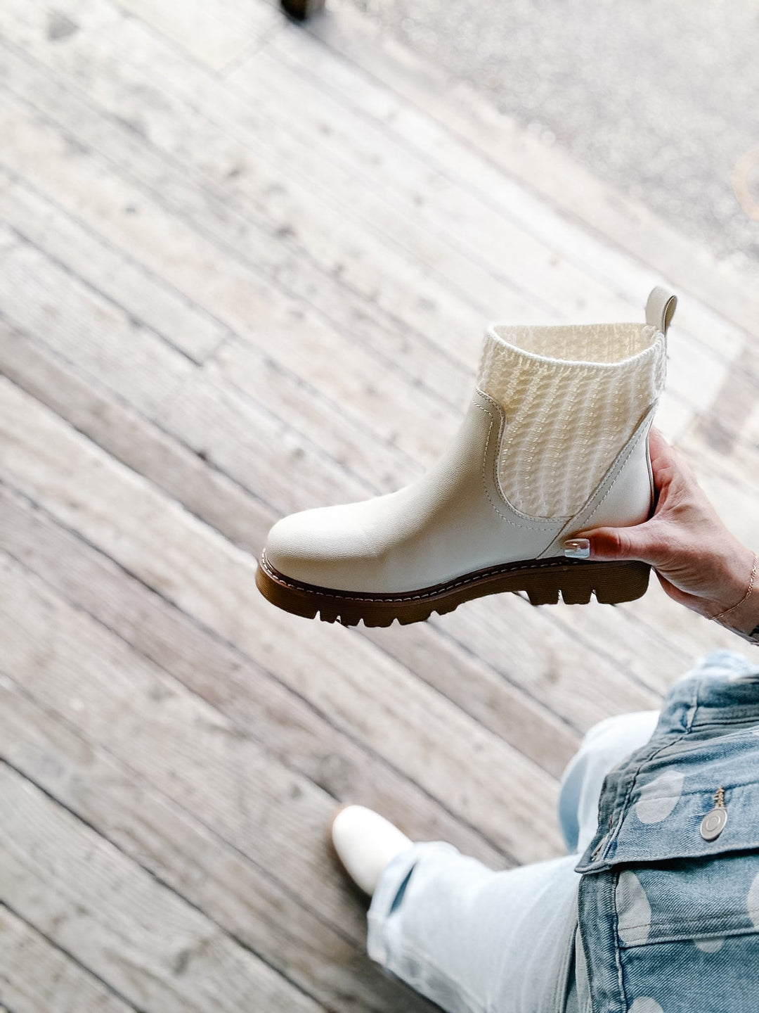A hand holds up a cream-colored Chelsea boot with a knit ankle cuff and a brown lug sole, set against a background of a person sitting on a wooden deck.