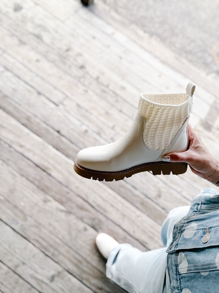 A hand holds up a cream-colored Chelsea boot with a knit ankle cuff and a brown lug sole, set against a background of a person sitting on a wooden deck.