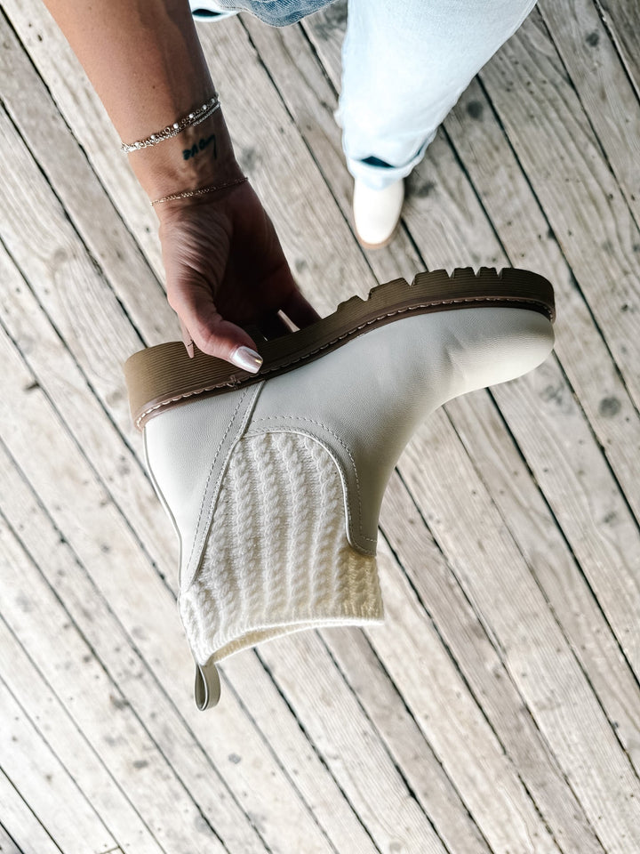 A hand holds up a cream-colored ankle boot with a textured knit side panel and a brown sole, set against a background of light-colored wooden planks with a person&
