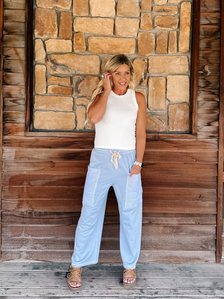 A woman stands on a wooden deck in front of a rustic stone and wood wall, wearing a white sleeveless top and light blue drawstring lounge pants.